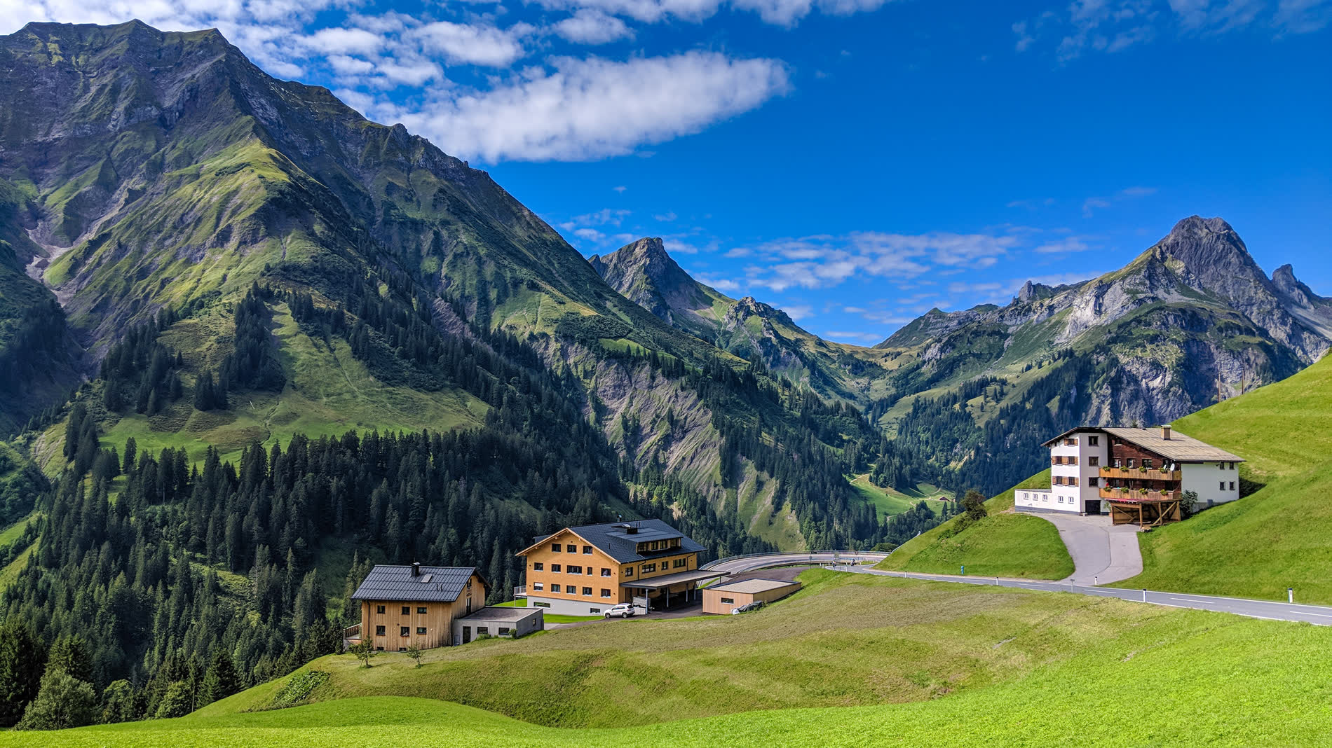 Bergtal mit Häusern und einer Straße, dahinter schroffe Gipfel unter blauem Himmel Bergtal mit Häusern und einer Straße, dahinter schroffe Gipfel unter blauem Himmel