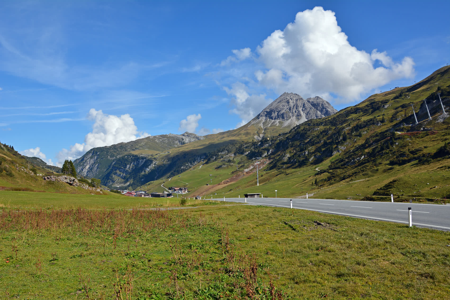 Passstraße durch grüne Almhänge, dahinter ein spitzer Felsgipfel unter Wolken Passstraße durch grüne Almhänge, dahinter ein spitzer Felsgipfel unter Wolken