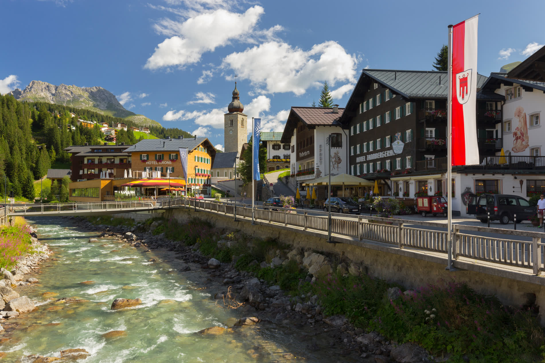 Gebirgsbach unter einer Uferstraße, dahinter Hotels, eine Kirche und eine rot-weiße Fahne Gebirgsbach unter einer Uferstraße, dahinter Hotels, eine Kirche und eine rot-weiße Fahne